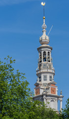 Fototapeta premium Amsterdam, the Netherlands - August 16, 2016: The spire of the Zuiderkerk against blue sky with green tree in lower left corner.