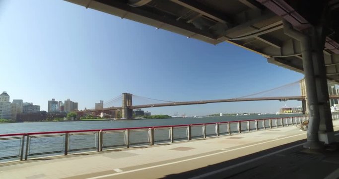 A dolly shot of people walking and jogging on the East River Bikeway in Manhattan with the Brooklyn Bridge in the background.  	
