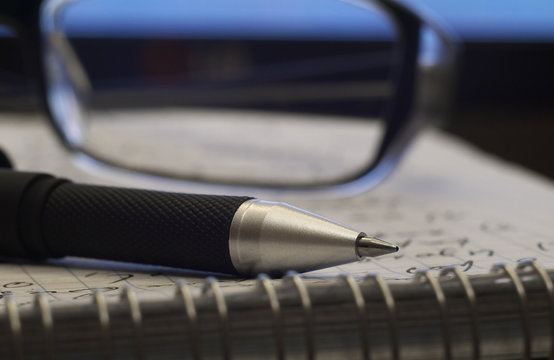 ballpoint pen on notebook closeup with glasses in background, shallow depth of field