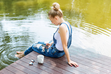 Young woman using her smartphone sitting on the pier and drinking hot coffee