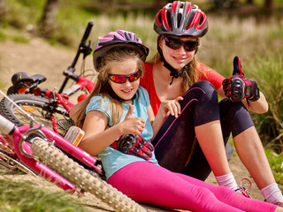 Happy mother and daughter are sitting on road near bicycles. Bicycle racing concept. Bicycle girl wearing helmet show thumb up.