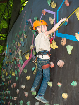 Little boy in helmet and with a safety rope climbing wall holding hooks on the blurred background of trees