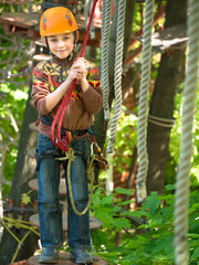Little boy in the helmet and a safety system stands on the suspension bridge and keeps the ropes against the sky and the trees with leaves
