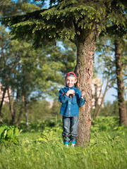 Little boy in a cap and a denim dress standing on the grass and keeps pine cones on blurred background forest