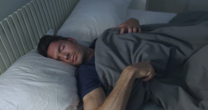 Sleeping man turns towards light while dreaming in a double bed with soft ambient light.  Camera move on jib arm.  Side view, medium shot.