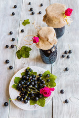 Preserved homemade black currant jam in glass jars on wooden table. Fresh berries and green leaves, vintage white plate, top view.