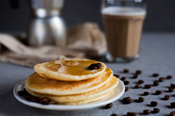 Pancakes decorated with coffee grains on a white plate, moka coffee maker on a vintage tablecloth, a glass of latte on a dark gray background, horizontal photo