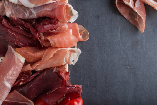 Jamon mix. Ham. Traditional Italian and Spanish salting, smoking, dry-cured dish - jambon Serrano and prosciutto crudo sliced with herbs and tomatoes on dark stone background. Copy space. Closeup. 