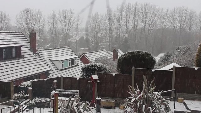 Snow falls on rear gardens and the roof tops of English, suburban houses. 