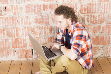 Handsome man typing on laptop in front of a brick wall. Hipster working in room. Modern lifestyle