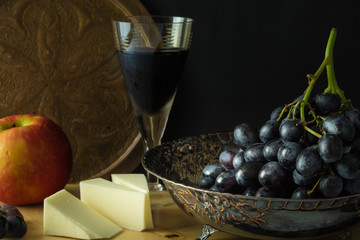 Antique fruit bowl with cluster of grapes, apple with pieces vegan cheese and a glass of red wine.
