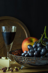 Antique fruit bowl with cluster of grapes, apple with pieces vegan cheese, pistachios and a glass of red wine