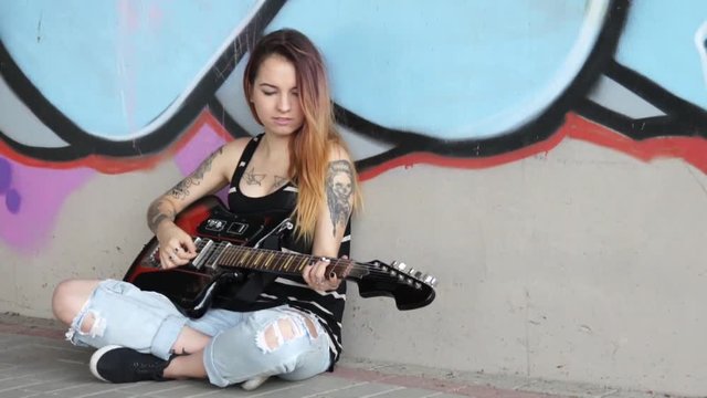 Girl sitting near a graffiti wall and plays electric guitar.