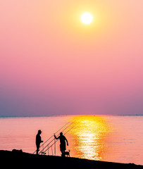 silhouette of a fisherman with a fishing rod on the shore at sunrise (sunset). Surface of sea water and the sun above the horizon.
