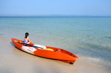young asian girl in swimming suit sitting on orange kayak with yellow paddle on a sandy beach