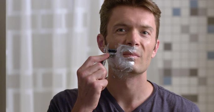 Man shaving with a wet razor and foam to remove stubble on cheek.  Slow motion recorded at 60fps, close up, front view from mirror POV.