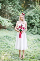 Captivating bride with red wedding bouquet stands in the grass