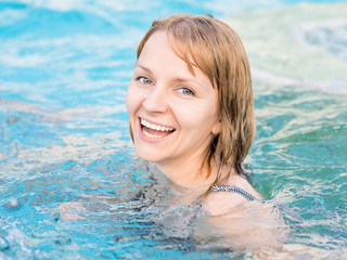 Smiling beautiful young woman in the swimming pool at aquapark. Portrait of a happy girl having fun enjoyable time on vacation. Looking at camera.