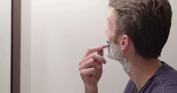 Man wet shaving to remove stubble while looking in a bathroom mirror.  Slow motion recorded at 60fps, close up, profile.