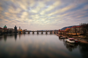 Fototapeta na wymiar Beautiful sunrise near famous Charles Bridge with cloudy sky and water reflection. Bridge with amazing tower and old statues at early morning. Typical sunny day in Prague, Czech republic.