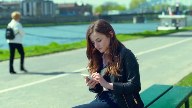 Pretty girl sitting on the bench next to the river and browsing internet on smartphone
