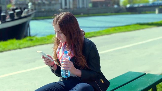 Girl drinking water on the bench and texting on smartphone
