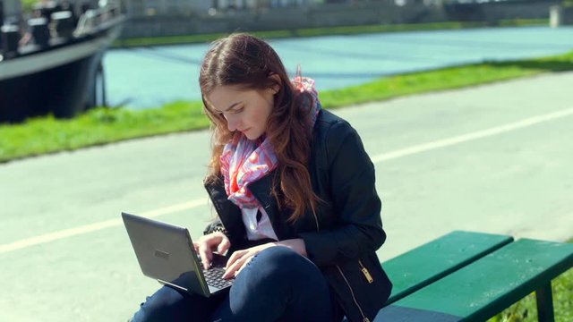 Girl sitting on the bench next to the river and using laptop
