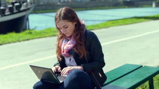 Girl answers cellphone while sitting on the bench and typing on laptop
