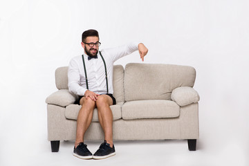 Picture of surprised handsome hipster man pointing down while sitting on sofa or couch isolated on white background in studio. Studio shot.