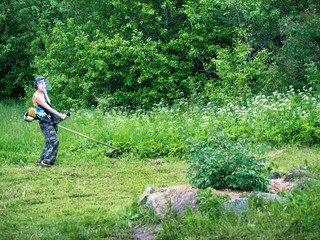 Adult woman in a protective mask holding a streamer and mowing grass against the backdrop of deciduous trees