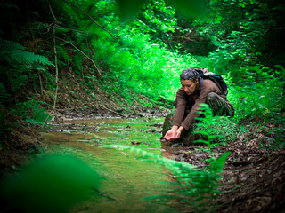 Toned image of an adult woman with a backpack and bandanna sitting on the bank of the stream and drinking water against the backdrop of dense forest with shrubs and ferns