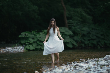 Fototapeta premium Fine art outdoor portrait of beautiful young woman in a white dress. Girl walking alone by the river.