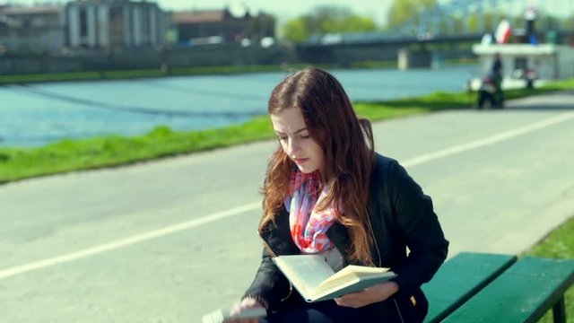 Pretty girl answers the phone while reading book on the bench
