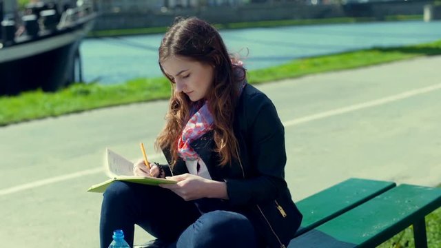 Girl writing something in her journal and drinking water on boulevard
