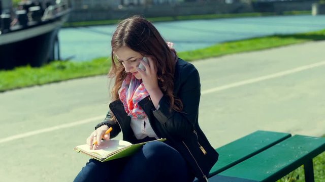 Girl talking on cellphone while writing in the journal on the bench
