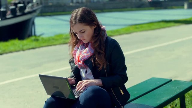 Girl using laptop while sitting on the bench and smiling to the camera
