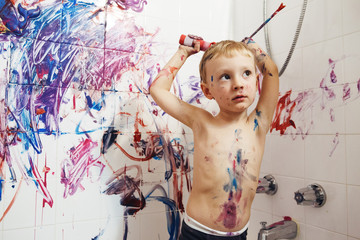 Fototapeta premium Portrait of cute adorable white Caucasian little boy playing and painting with paints on wall in bathroom having fun, lifestyle childhood concept