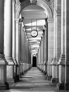 Fototapeta Długi korytarz z kolumnami i zegarem zwisającym z sufitu. Perspektywa krużgankowa. . Obraz czarno-biały.