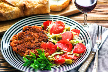 Homemade Breaded German Weiner Schnitzel and fresh vegetable spring salad with tomato, green olives, cabbage and parsley in white bowl on wooden table, closeup.