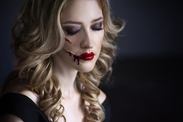 Beautiful young blonde woman in black dress with halloween make up and bloody face art, victim of...
