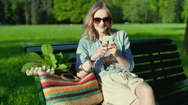 Pretty girl sitting on the bench in the park and texting on smartphone
