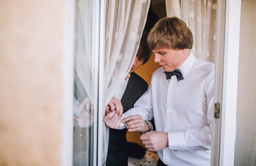 Fototapeta premium Groom and his friends prepare for the wedding. Grooms man helps the groom to put on black cufflinks on white sleeve of the shirt. Details of the morning of the wedding day