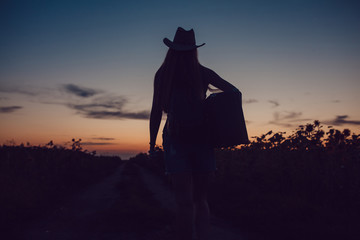 Girl in a cowboy hat standing with a suitcase on the road in the sunflower field. Waiting for help. Sunset.