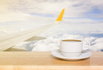Wooden table and cup coffee with blurred airport as seen through window of an aircraft after takeoff. Wing of the plane on airport in background