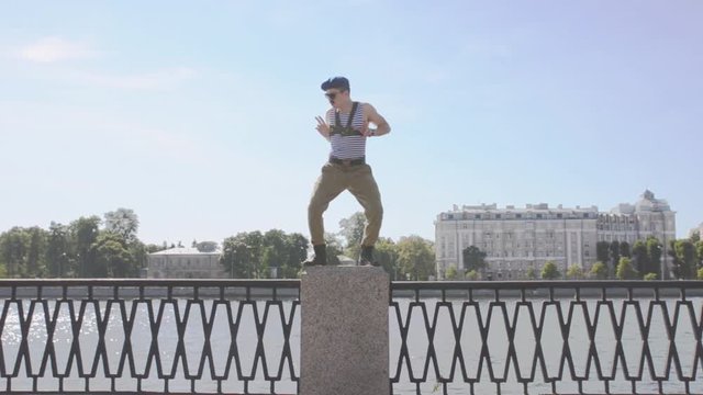 Man in marine uniform, sunglasses show weird gesture, dance staying on postament at seafront. Surrealism. Water, summer sunny day