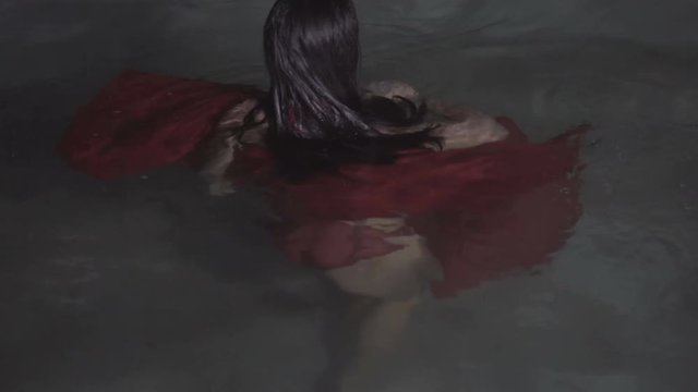 Closeup portrait of beautiful mysterious woman wearing red dress looking into the camera while dancing in the pool at night