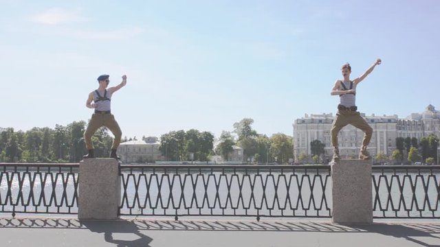 Two men in marine uniform show weird movements, staying on postament at seafront. Surrealism. Water, summer sunny day