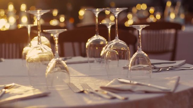 Slow motion shooting, empty wine glasses on the table with white tablecloth, sunshine, Italian summer evening, elegant atmosphere of wedding. Camera stabilizer shot.