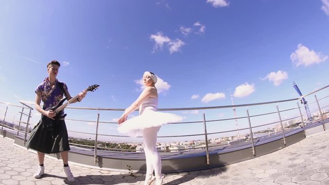 Man in skirt play electric guitar on seafront, sing in camera. Girl dance in ballerina suit. Crazy style