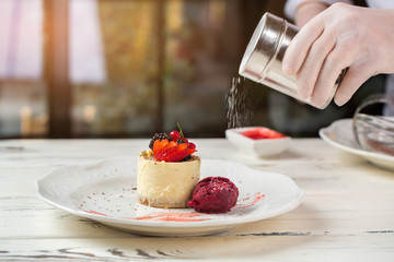 Powder falls on ice cream. Plate with little cake. Cheesecake and powdered sugar. Dessert is served cold.